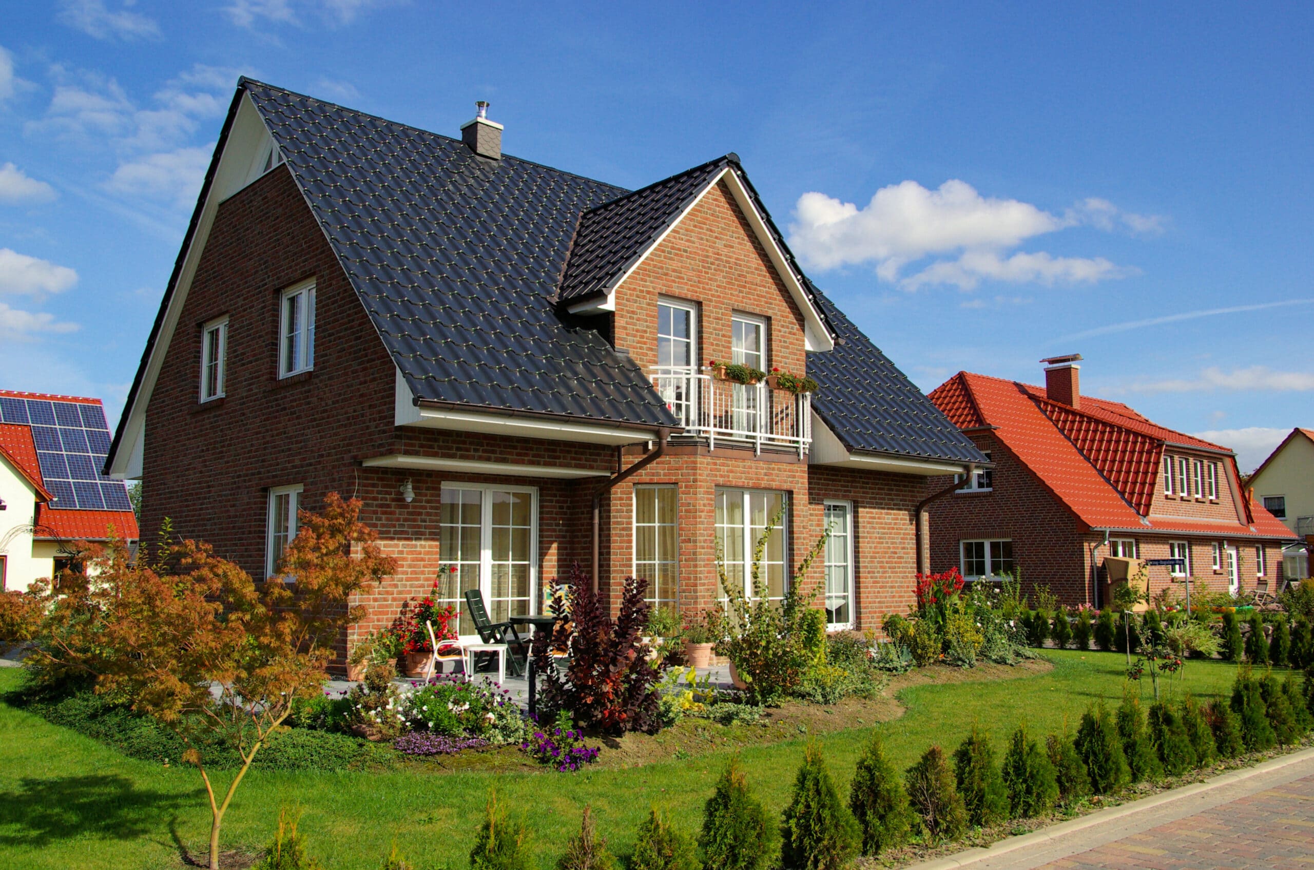 Modern brick house with dark tiled roof and small balcony, blue sky above, and a well-kept front garden with flowering plants.
